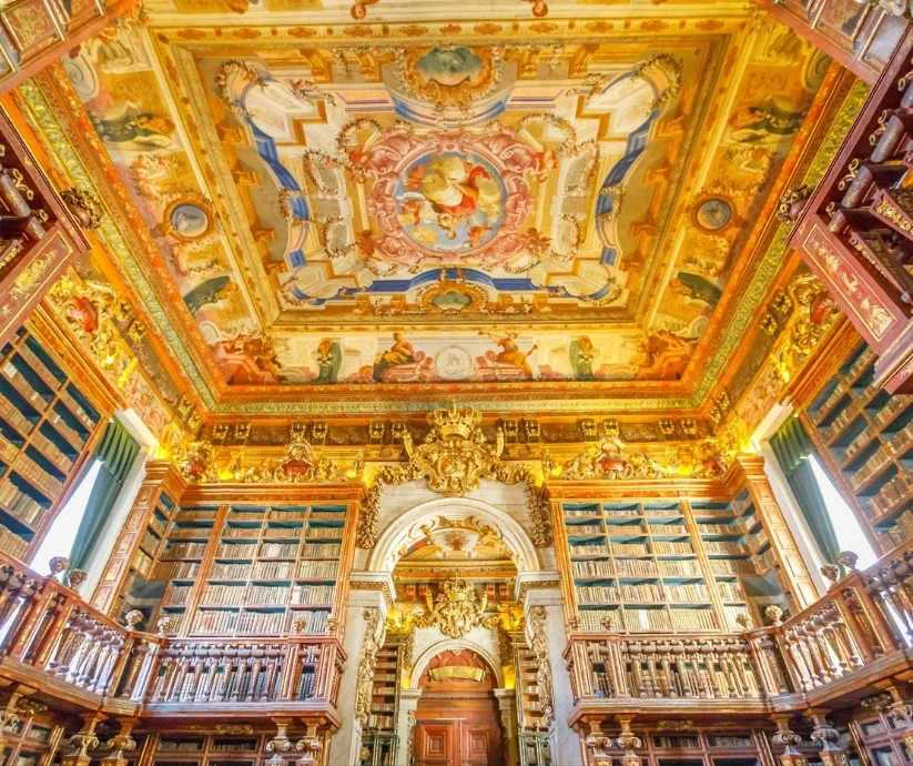 Interior de la Biblioteca Joanina en Coímbra, Portugal, con estanterías de madera, techos decorados y detalles dorados.