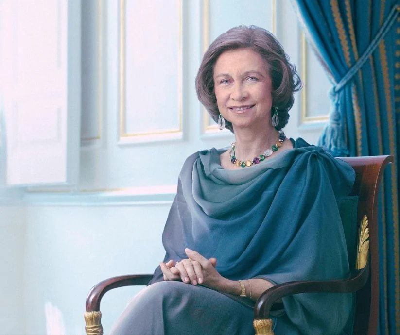 Retrato oficial de la reina Sofía de España, sentada en un salón palaciego, con vestido gris y joyas de colores.